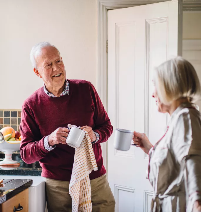 Image of a cheerful senior man in a burgundy sweater enjoying coffee in a kitchen while conversing with a woman, representing personalized retirement lifestyle content.