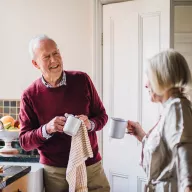 Image of a cheerful senior man in a burgundy sweater enjoying coffee in a kitchen while conversing with a woman, representing personalized retirement lifestyle content.