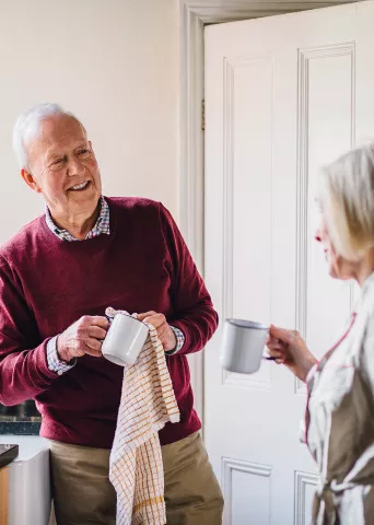 Image of a cheerful senior man in a burgundy sweater enjoying coffee in a kitchen while conversing with a woman, representing personalized retirement lifestyle content.