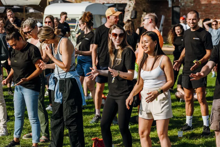 A diverse group of people joyfully dance at the Springbok summer team event on a sunny day. They are casually dressed, conveying a relaxed and festive atmosphere.