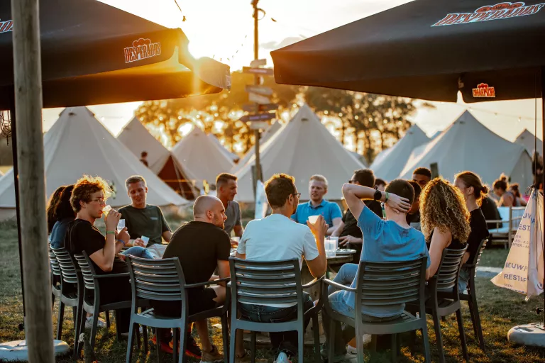 A group of people sit around a table, enjoying drinks under large umbrellas at sunset, surrounded by tents and trees at the Springbok summer team event.