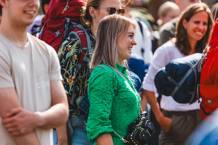 A woman from Springbok Agency in a vibrant green shirt smiles amidst a crowd, with backpacks visible, suggesting an outdoor event. The atmosphere is lively and cheerful.