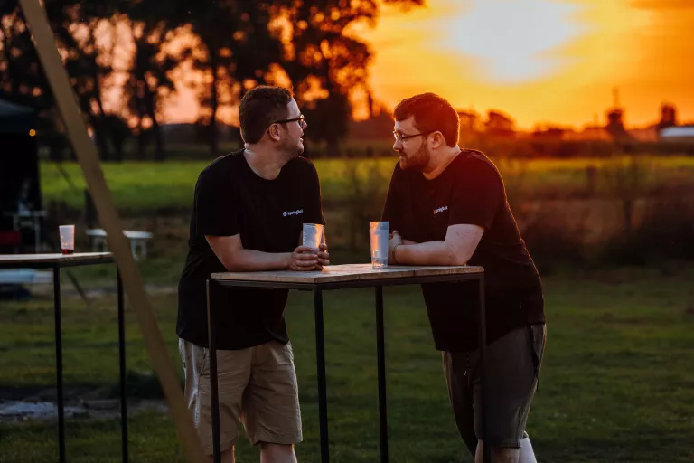 Two men in black shirts lean on a high table, holding cups, conversing at sunset at Springbok's summer team event.