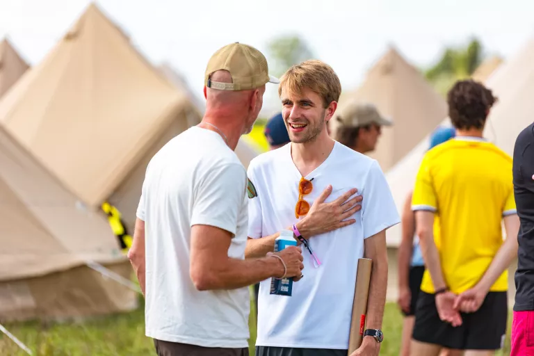 Two men engage in friendly conversation at Springbok's outdoor team event with beige tents in the background. One wears a white shirt, smiling warmly, while the other in a cap holds a drink. 