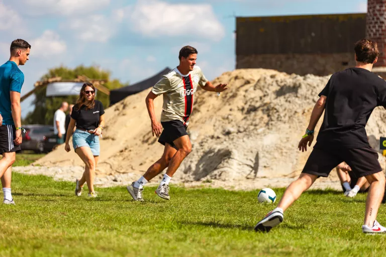 A group of Springbok employees plays soccer on a grassy field, with a woman and three men actively engaged. A sandy mound and cloudy sky form the background.