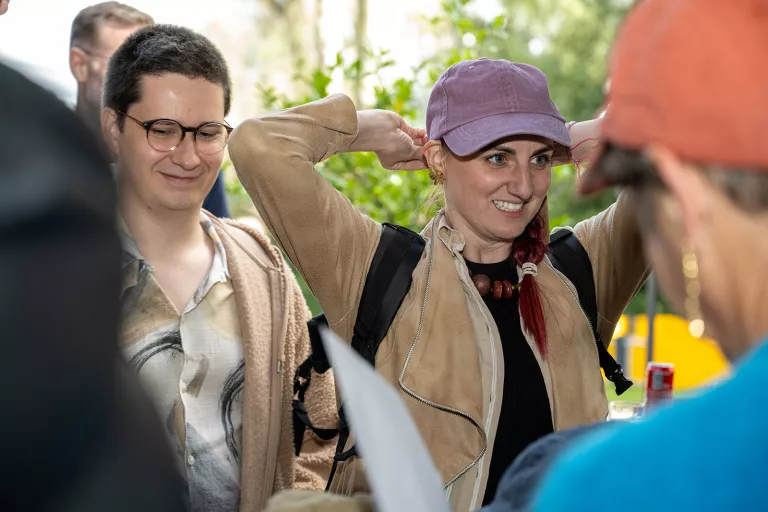 A woman in a purple cap smiles, adjusting her hair, standing beside a man in glasses. They are outdoors at the Springbok summer team event, surrounded by greenery and people.