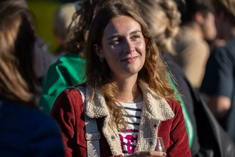 A woman from the Springbok Agency team with wavy brown hair smiles softly in a sunlit outdoor gathering, wearing a red corduroy jacket and holding a drink. The mood is relaxed and joyful.