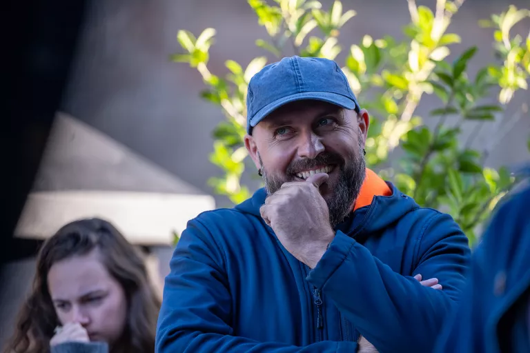Man in a blue cap and hoodie smiles thoughtfully with his hand on chin, standing outside near greenery, listing to a speech.
