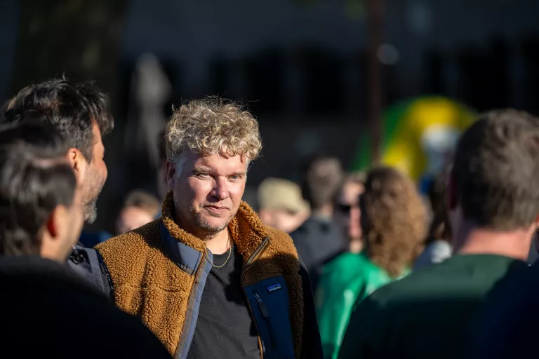 A curly-haired person in a brown fleece jacket at Springbok's outdoor team event. The mood is casual and social, with blurred people in the background.