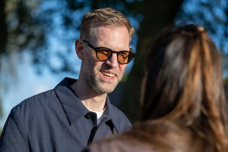 Man with sunglasses smiling in conversation, wearing a navy shirt. His expression is friendly, with sunlight filtering through trees.