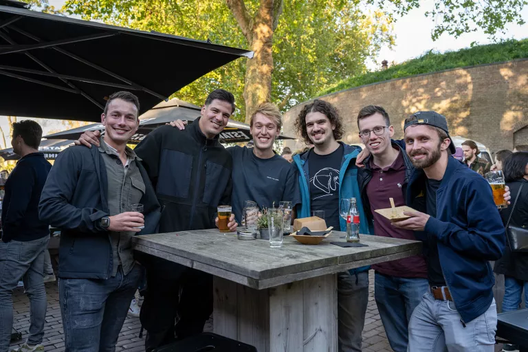 A group of six smiling Springbok people stand around a rustic outdoor table with drinks in hand. The mood is cheerful and relaxed under dappled sunlight and leafy trees.