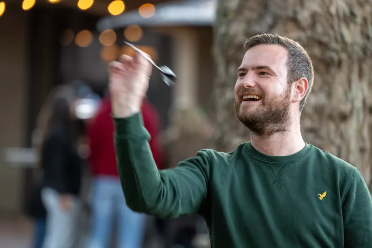 A bearded man in a green sweater joyfully throws a dart outdoors, with a blurred background of people and warm string lights, conveying a lively mood.