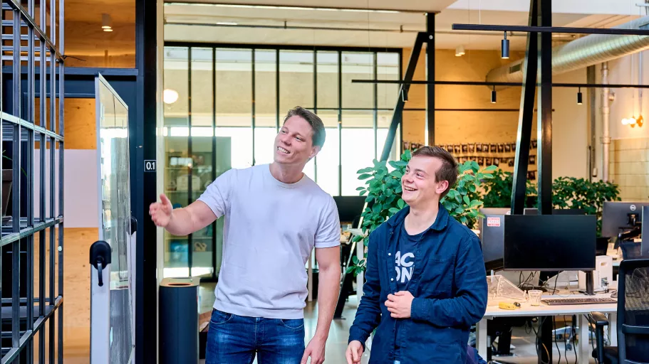 Two Springbok team members smiling and gesturing while walking through a modern office with large windows and desks. The atmosphere is friendly and casual.