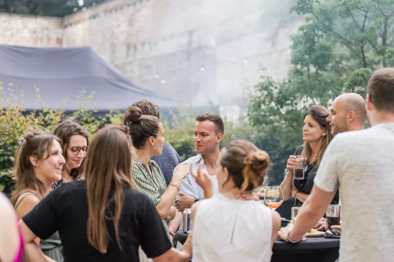 A group of people engage in animated conversation at Springbok's summer barbecue event in Ghent. The setting is relaxed, with trees and a brick wall in the background.