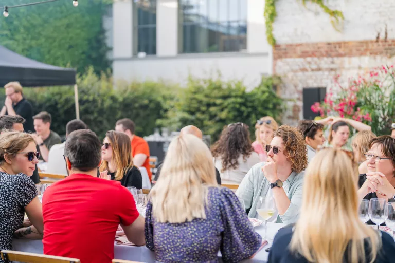 People seated at tables during the Springbok summer barbecue in Ghent, enjoying drinks and conversation. The setting is relaxed, with greenery and a brick wall backdrop.