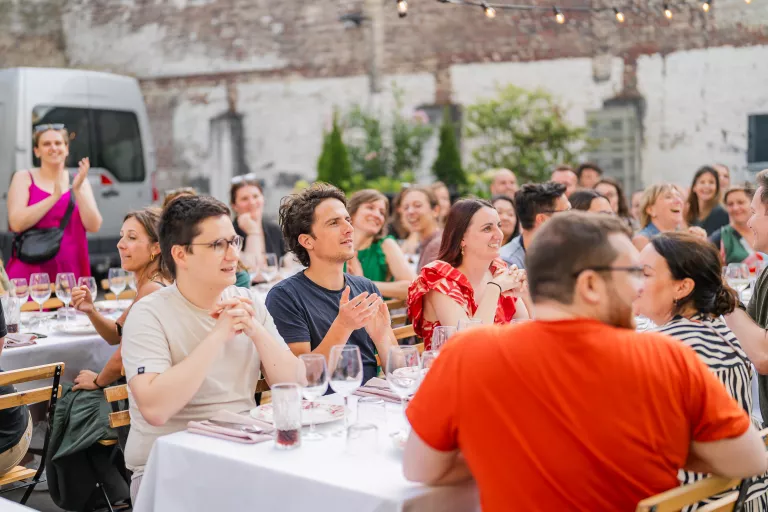 A cheerful Springbok summer barbecue gathering in Ghent with people seated at tables, applauding. String lights hang above, creating a festive and joyful atmosphere.