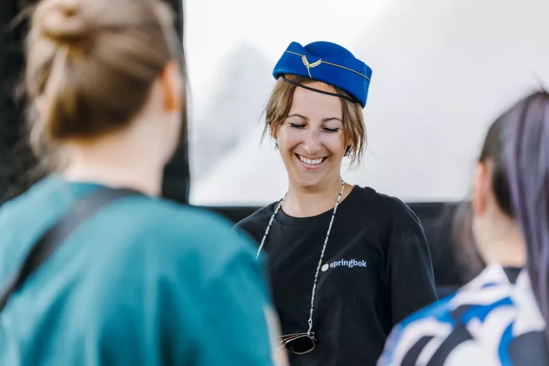 Smiling woman in a blue hat, wearing a "Springbok" shirt, engages with two blurred people in the foreground, conveying a friendly atmosphere.