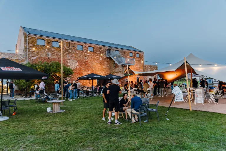 Outdoor Springbok event at dusk near a brick building with string lights. Groups gather under tents and umbrellas on a grass lawn, creating a lively, social atmosphere.