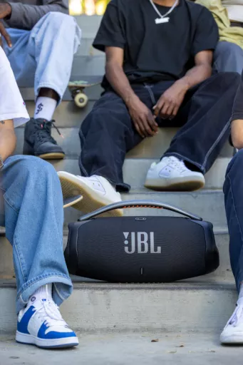 Young people sit casually on steps next to a large JBL speaker, conveying a relaxed, social atmosphere. Skateboard visible, wearing casual jeans and sneakers.