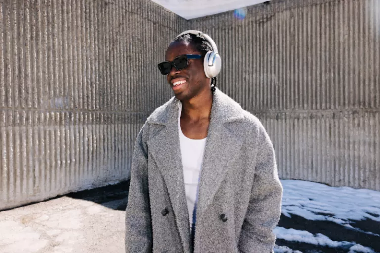 Smiling man wearing headphones, sunglasses, white shirt, and a gray winter coat stands outdoors on a snowy pavement, with textured concrete walls.